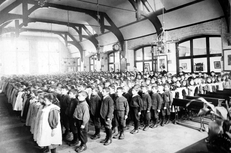 Children stand to attention in lines for an assembly in the Central Hall at Darley Street School, with teachers at the back.