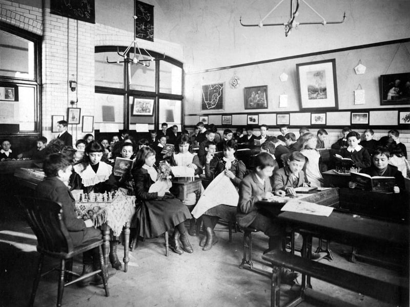 Children pursuing their hobbies, which include chess, reading and sewing. A girl in the centre is nursing a doll. The girls are wearing boots with thick stockings.