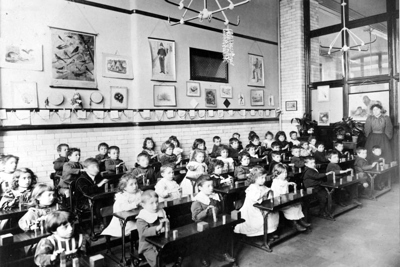 A class of infants learning to build with toy bricks. The children are sitting at their desks, each with a small amount of bricks. The teacher, on the right, is wearing a blouse with 'leg-o-mutton' sleeves.