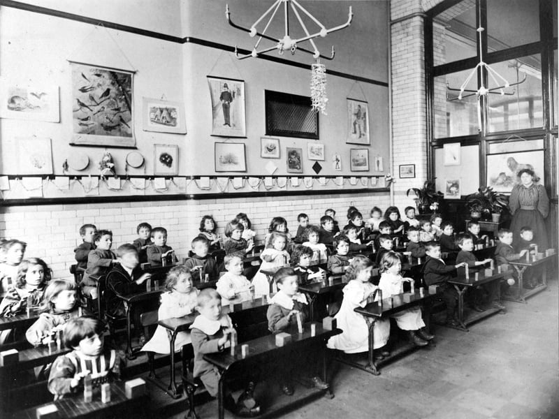 A class of infants learning to build with toy bricks. The children are sitting at their desks, each with a small amount of bricks. The teacher, on the right, is wearing a blouse with 'leg-o-mutton' sleeves.