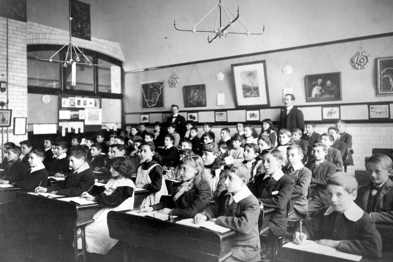 Girls and boys are seated two to a desk with two schoolmasters standing behind. The children are writing in their books, perhaps copying information from the chalk board.