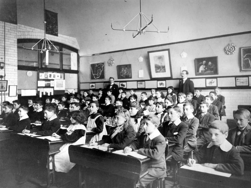Girls and boys are seated two to a desk with two schoolmasters standing behind. The children are writing in their books, perhaps copying information from the chalk board.