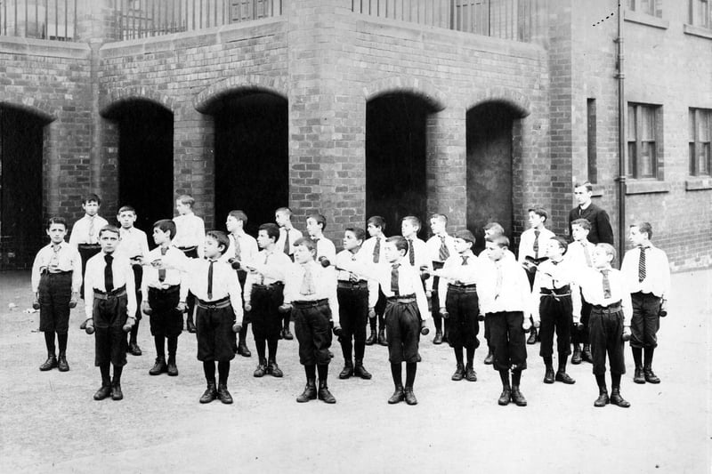 Class of boys in school playground having a drill lesson, the fore-runner of physical education. Each boy has a weight in each hand, in the manner of regimental drill, movements would be made in syncronized fashion.