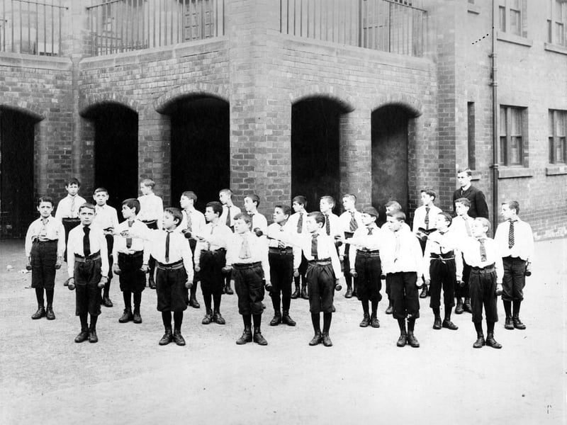Class of boys in school playground having a drill lesson, the fore-runner of physical education. Each boy has a weight in each hand, in the manner of regimental drill, movements would be made in syncronized fashion.