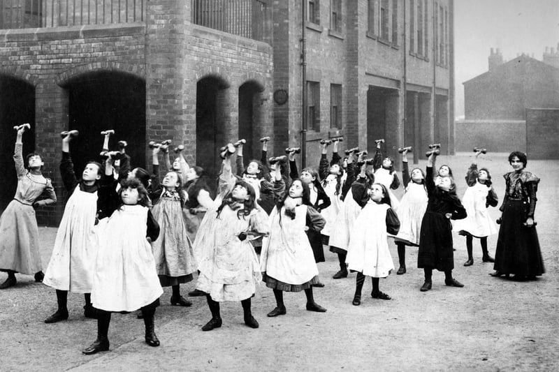 Class of girls having a drill lesson in the playground at Darley Street School. Each girl has a weight in each hand, rhythmic movements are made on command of the teacher.